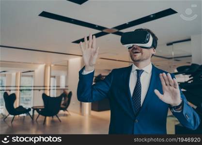 Businessman in futuristic VR headset touches air with his palms immersed in virtual reality. Young financier using innovative method of gaming experience standing in office lobby during coffee break. Businessman in futuristic VR headset touches air with his palms immersed in virtual reality