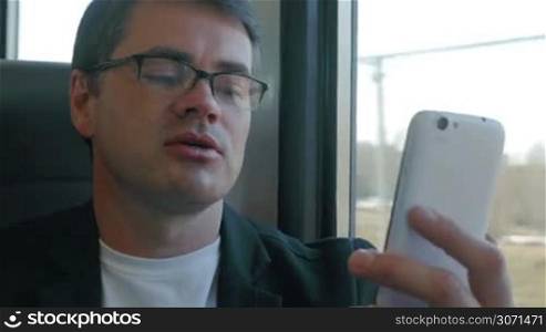 Businessman in glasses talks cheerfully on skype using smartphone while travelling by train