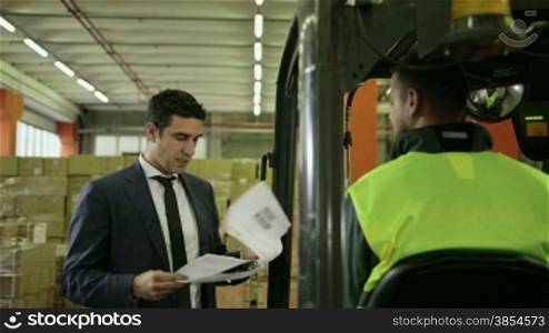 Businessman in shipping facility speaking with manual worker operating forklift, people working in warehouse, workers in industry. 10of19