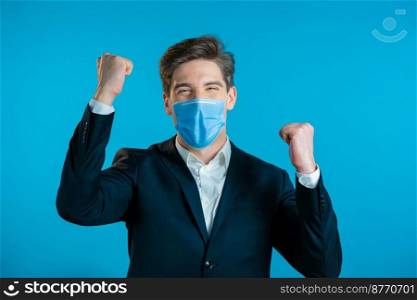 Businessman in suit is very glad and happy, he shows yes gesture of victory, guy achieved result, goals. Surprised excited happy guy on blue background.. Businessman in suit and medical mask is very glad and happy, he shows yes gesture of victory, guy achieved result, goals. Surprised excited happy guy on blue background