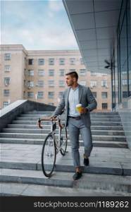 Businessman in suit with bicycle comes down the stairs at the office building in downtown. Business person riding on eco transport on city street, urban style. Businessman with bicycle comes down the stairs