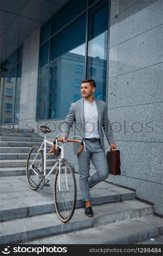 Businessman in suit with bicycle comes down the stairs at the office building in downtown. Business person riding on eco transport on city street, urban style