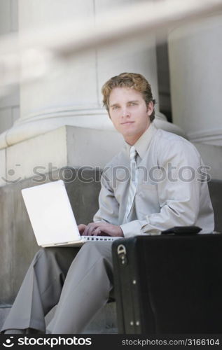 businessman in tan shirt and pants is typing on his white laptop on the steps of a courthouse, with a black briefcase in the foreground