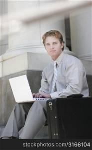 businessman in tan shirt and pants is typing on his white laptop on the steps of a courthouse, with a black briefcase in the foreground