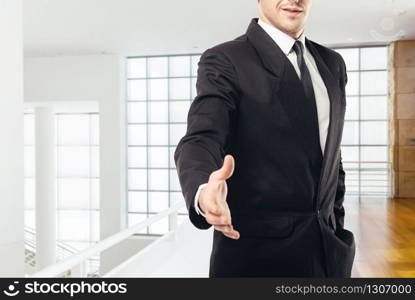 Businessman in tie and black suit extends his hand, business center with glass walls on background
