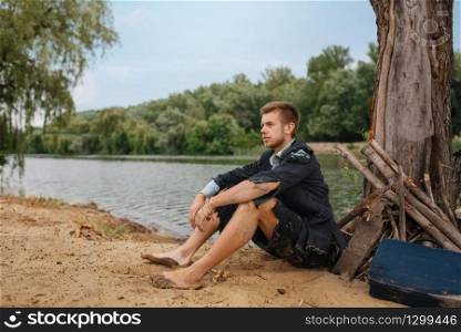Businessman in torn suit sitting on the sand on lost island. Business risk, collapse or bankruptcy concept