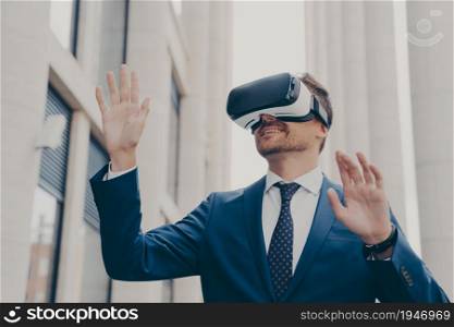 Businessman in virtual reality glasses using VR goggles while standing on city street outdoors, testing innovative method for business, gesturing while interacting with digital interface. Businessman in virtual reality glasses using VR goggles while standing on city street outdoors