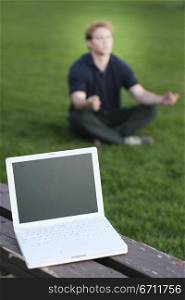 Businessman is meditating in the green grass as his white laptop sits on the brown bench