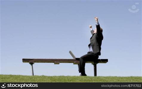 Businessman is waving his hands in celebration as he sits on a park bench, next to his laptop