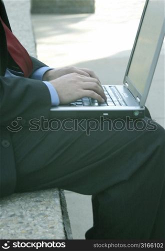 Businessman is wearing a red tie and is typing on his laptop outside on a bench