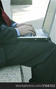 Businessman is wearing a red tie and is typing on his laptop outside on a bench