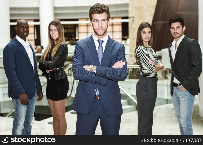 Businessman leader looking at camera with arms crossed in office building. Group of muti-ethnic people in the background