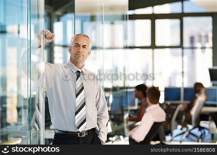 Businessman leaning on door in office with office workers in background.