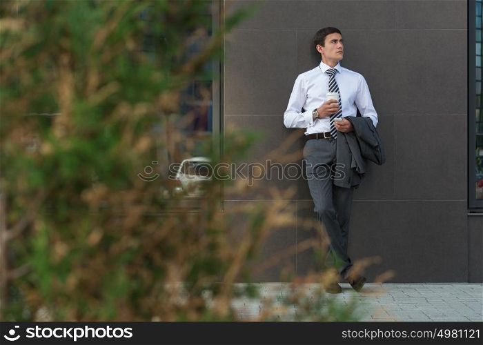 Businessman Leaning on Modern Office Building Wall With Coffee Using Mobile Phone