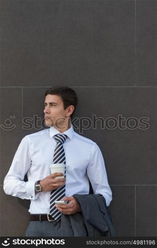 Businessman Leaning on Modern Office Building Wall With Coffee Using Mobile Phone