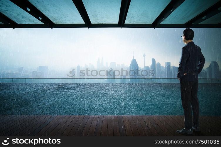 Businessman looking and thinking front of a Swimming pool on roof top with beautiful city skyline view,rainingday .