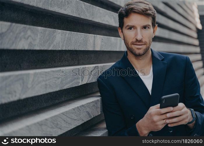 Businessman looks thoughtfully into distance uses smartphone thinks about future career success holds mobile phone wears formal elegant clothes stands near wall. Male employee with modern gadget. Businessman looks thoughtfully into distance uses smartphone thinks about future career success