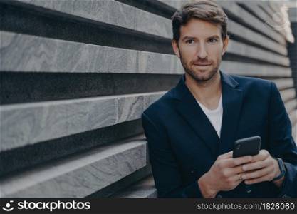 Businessman looks thoughtfully into distance uses smartphone thinks about future career success holds mobile phone wears formal elegant clothes stands near wall. Male employee with modern gadget. Businessman looks thoughtfully into distance uses smartphone thinks about future career success