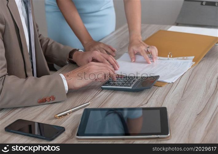Businessman manager in brown suit using calculator while sitting at office wooden desk and discussing on business document with female colleague in blue dress. Business analyst, investment, accounting, finance and teamwork concept.