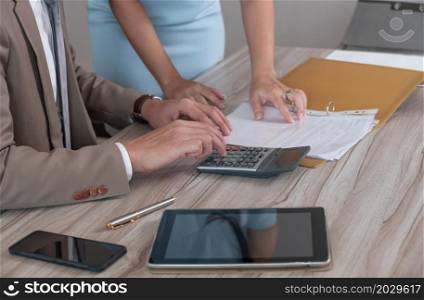 Businessman manager in brown suit using calculator while sitting at office wooden desk and discussing on business document with female colleague in blue dress. Business analyst, investment, accounting, finance and teamwork concept.