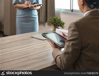 Businessman manager in brown suit using PC tablet while sitting at office desk and secretary in blue dress taking notes while standing in front of his boss. Business work concept