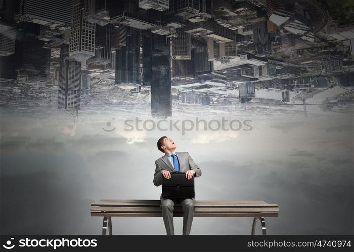 Businessman on bench. Young businessman sitting on bench with briefcase in hands and city reflecting in sky