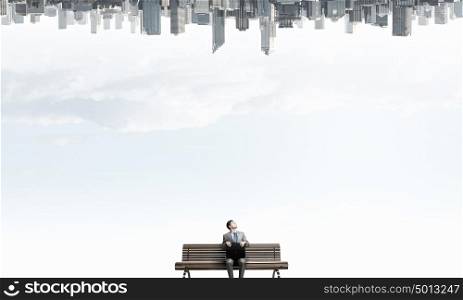 Businessman on bench. Young businessman sitting on bench with briefcase in hands and city reflecting in sky