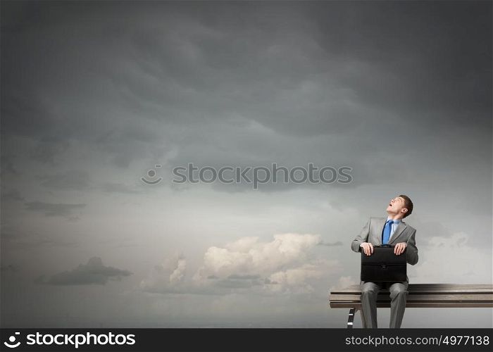 Businessman on bench. Young scared businessman sitting on bench with briefcase in hands and looking up