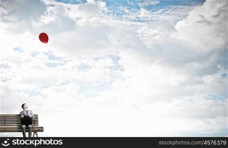 Businessman on bench. Young smiling businessman sitting on bench with briefcase in hands and looking at balloon in sky