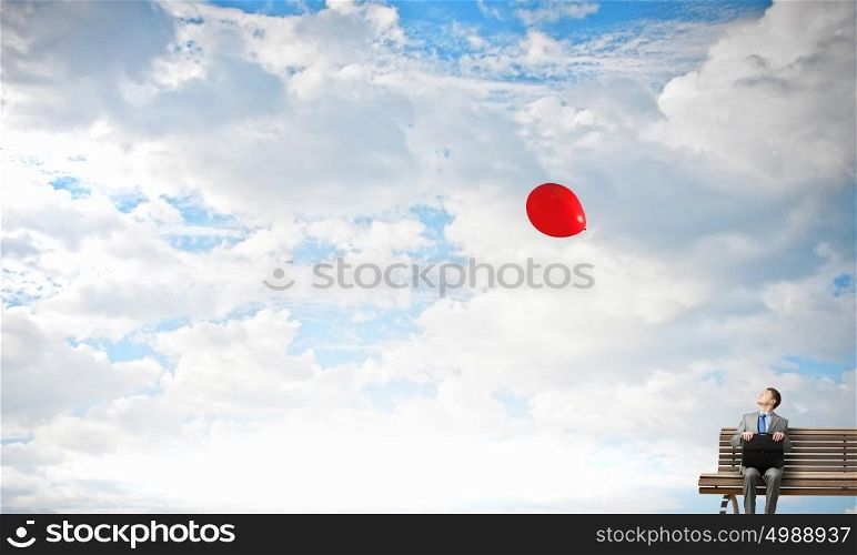 Businessman on bench. Young smiling businessman sitting on bench with briefcase in hands and looking at balloon in sky