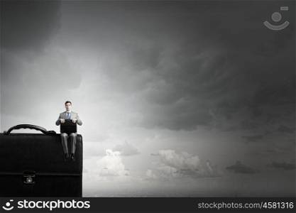 Businessman on big suitcase. Young smiling businessman sitting on giant briefcase