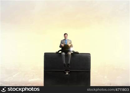 Businessman on big suitcase. Young smiling businessman sitting on giant briefcase
