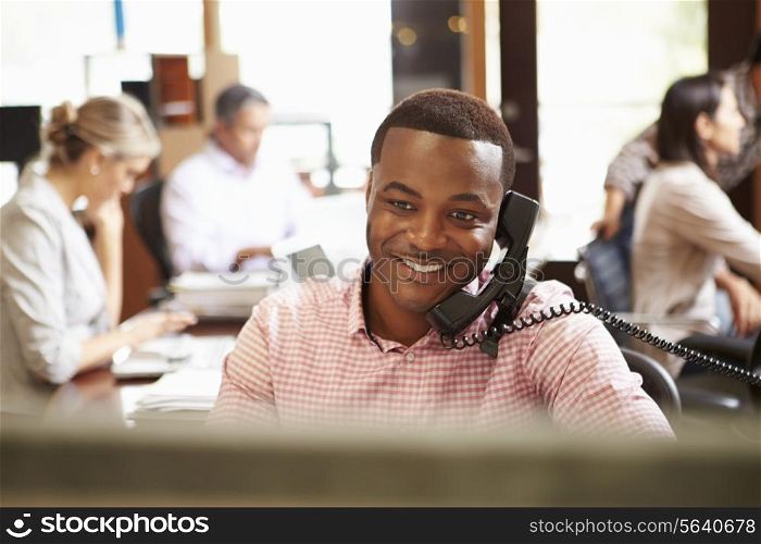 Businessman On Phone At Desk With Meeting In Background