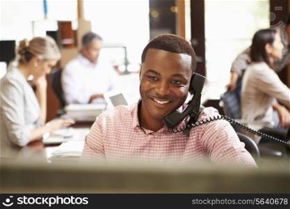Businessman On Phone At Desk With Meeting In Background