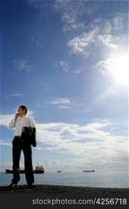 Businessman on the phone standing by the ocean with tankers in the background