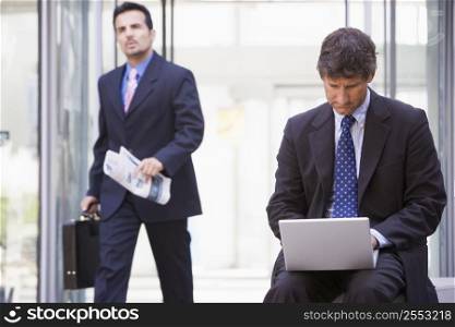 Businessman outdoors in front of building using laptop with businessman in background carrying newspaper (high key/selective focus)