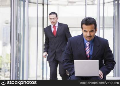 Businessman outdoors in front of building using laptop with businessman in background (high key/selective focus)