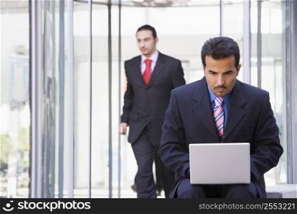 Businessman outdoors in front of building using laptop with businessman in background (high key/selective focus)