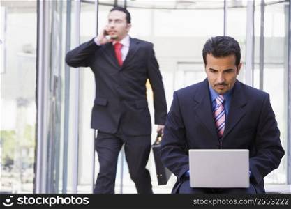 Businessman outdoors in front of building using laptop with businessman in background on cellular phone (high key/selective focus)