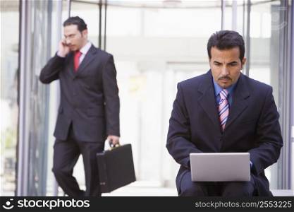 Businessman outdoors in front of building using laptop with businessman in background on cellular phone (high key/selective focus)