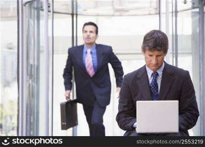 Businessman outdoors in front of building using laptop with businessman running in background (high key/selective focus)