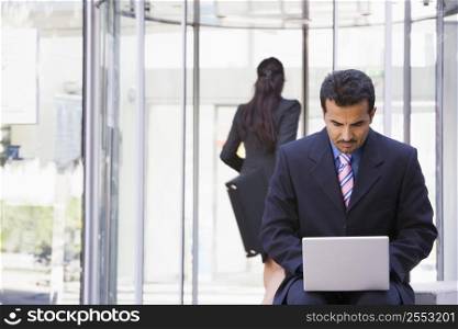 Businessman outdoors in front of building using laptop with businesswoman in background (high key/selective focus)