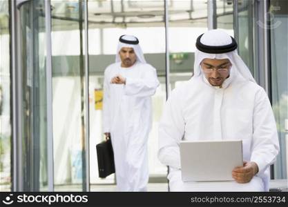 Businessman outdoors using laptop with businessman walking in background checking watch (selective focus)