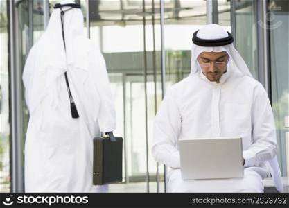 Businessman outdoors using laptop with businessman walking in background (selective focus)