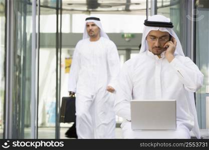 Businessman outdoors with laptop on cellular phone with businessman walking in background (selective focus)