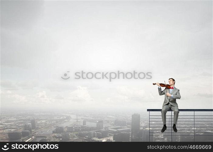 Businessman play violin. Young man in suit sitting on parapet and playing violin