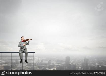 Businessman play violin. Young man in suit sitting on parapet and playing violin