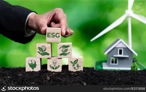 Businessman put ESG symbol cube with house and wind turbine model. Concept for forest regeneration by ethical corporate company with environmental awareness for green environment in future. Alter. Businessman put ESG symbol cube with house and wind turbine model. Alter