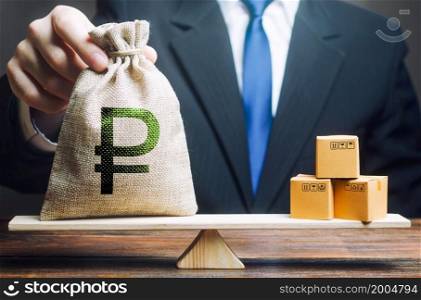 Businessman puts a russian ruble money bag and bunch of boxes on scales. Trade exchange balance. Retail distribution. Trade balance. Import export, economic processes. Profit from manufactured goods.