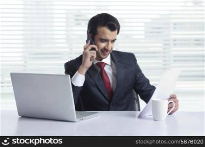 Businessman reading document while answering mobile phone at office desk